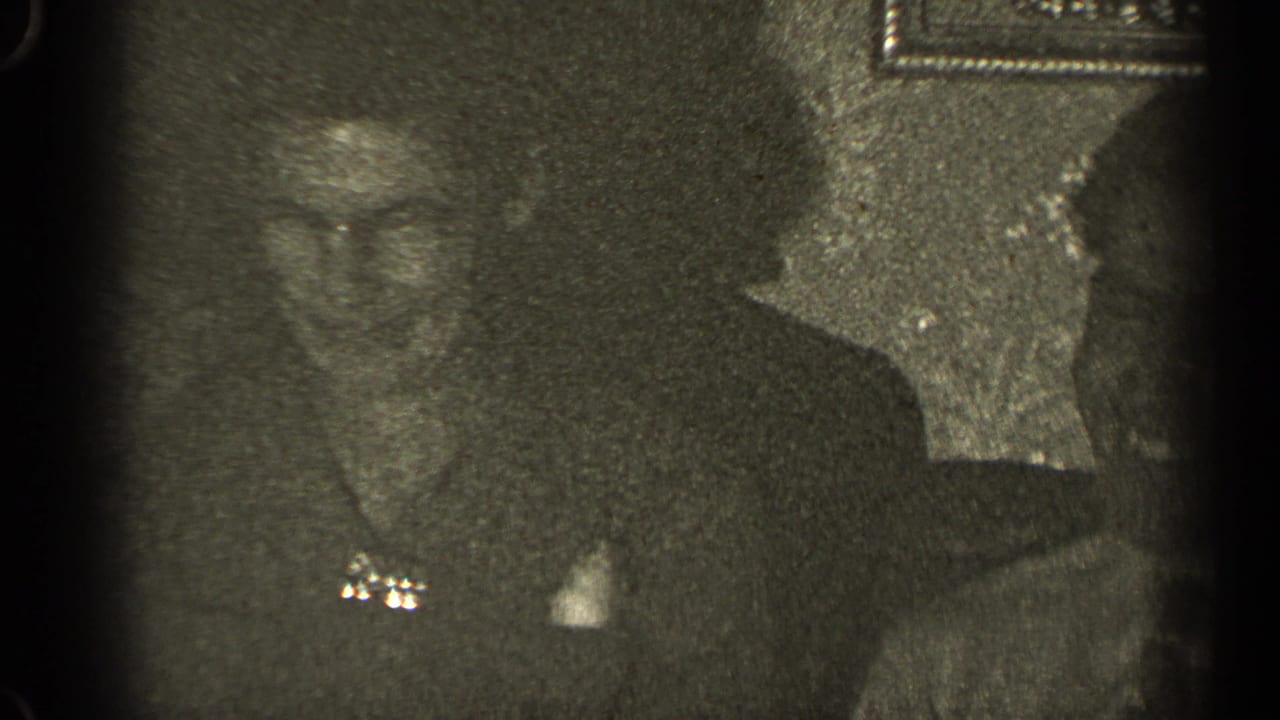 Adults in dark clothing sit close together in a dim indoor home-movie frame.