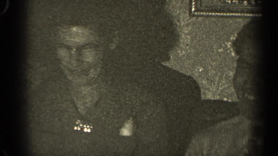 1947 Illinois 8mm frame showing adults in dark formal clothing seated indoors, with soft focus and visible film grain.