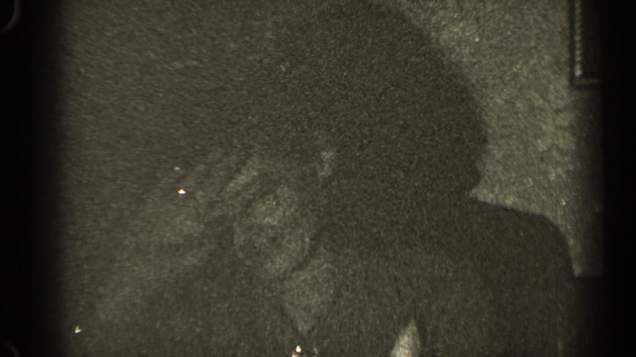 Grainy 1947 Illinois home-movie frame with adults seated indoors, one figure turned in profile during conversation.