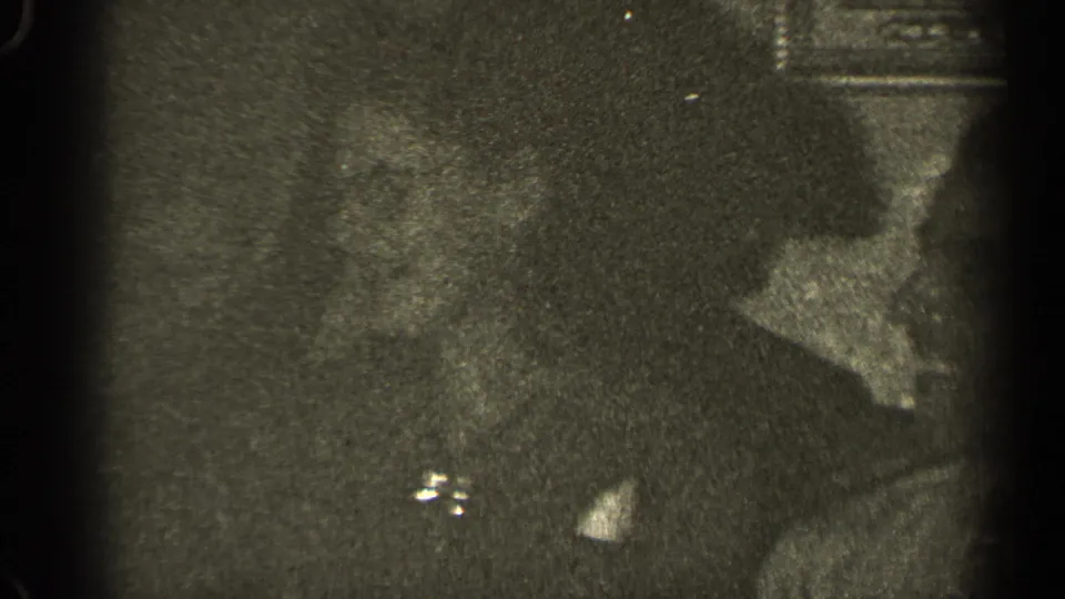 1947 Illinois home-movie frame showing adults gathered indoors, their faces soft and partly shadowed beside a wall decoration.
