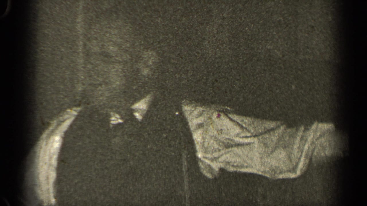 Partial adult figures in dark and light clothing seen in a grainy 8mm frame.