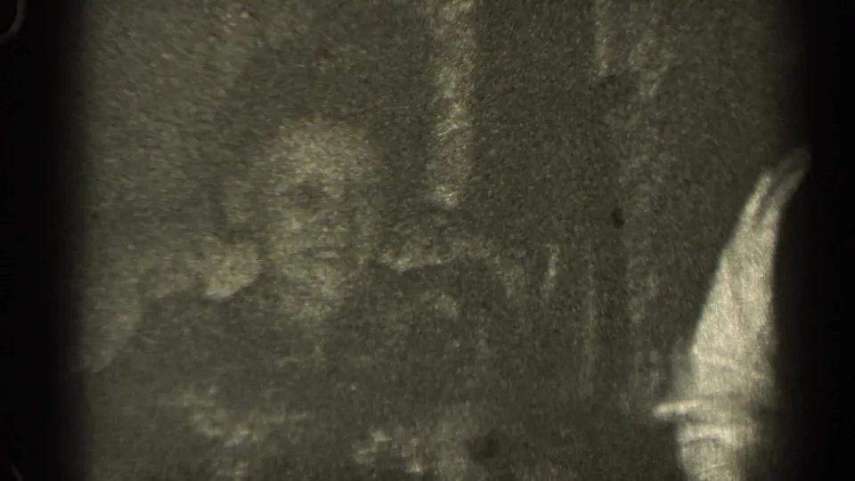 1947 Illinois home-movie frame with a partial adult in dark clothing beside a light sleeve, rendered in grainy black-and-white film.