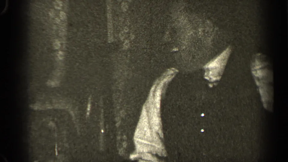 Adult at a 1947 Illinois indoor gathering, partly cropped, wearing dark clothing in grainy black-and-white 8mm footage.