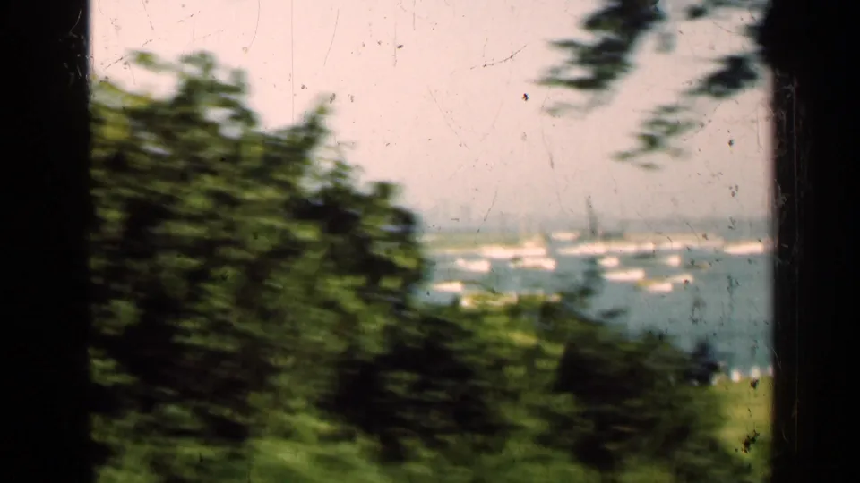 1947 Illinois 8mm frame looking past trees toward water with scattered boats and distant vessel shapes under a pale sky.