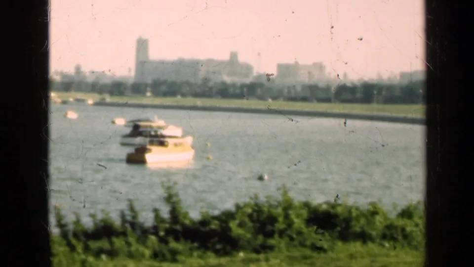 1947 Illinois waterfront frame with a small boat near the foreground, green shoreline, and hazy city buildings across the water.