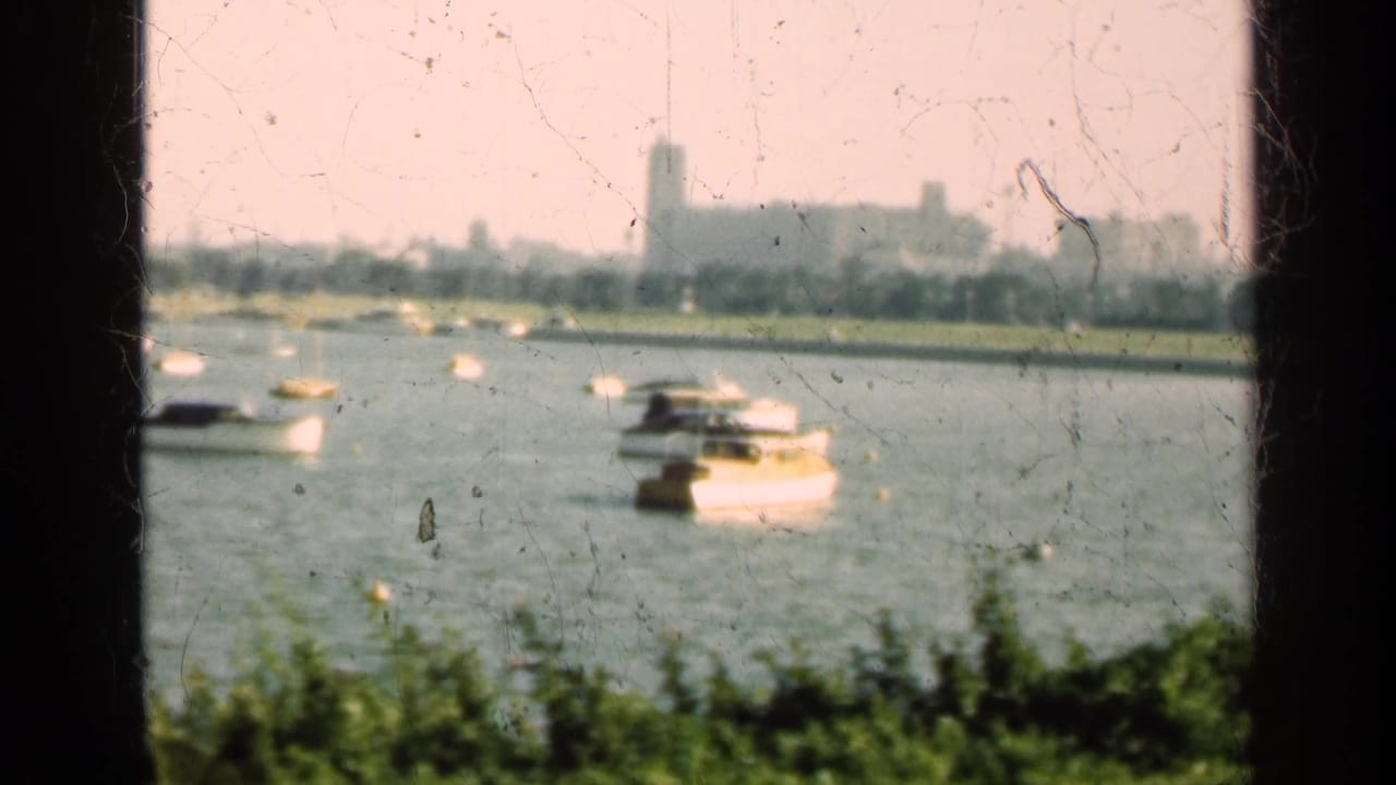 1947 Illinois 8mm view of white boats on a lake, with shoreline buildings and a tower faintly visible beyond dark frame bars.