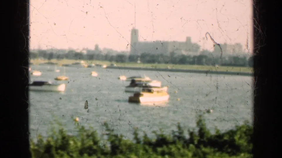 1947 Illinois 8mm view of white boats on a lake, with shoreline buildings and a tower faintly visible beyond dark frame bars.