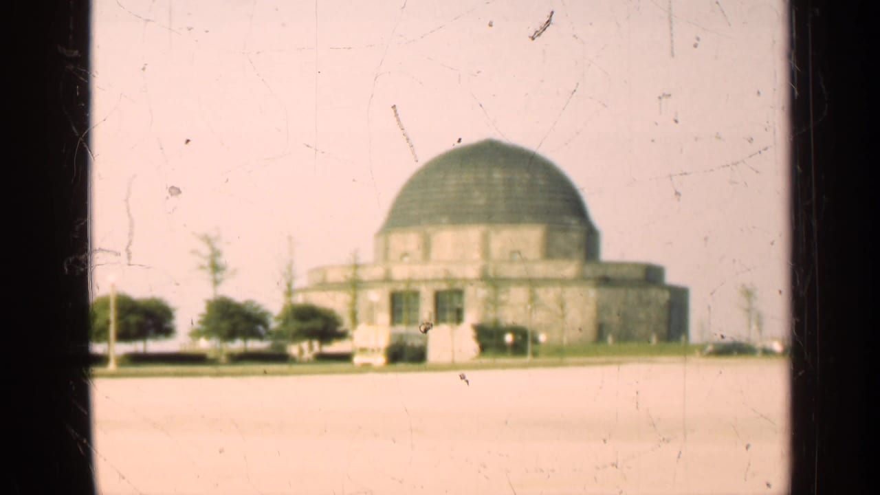 Large domed building with trees and cars in front, viewed across pavement.