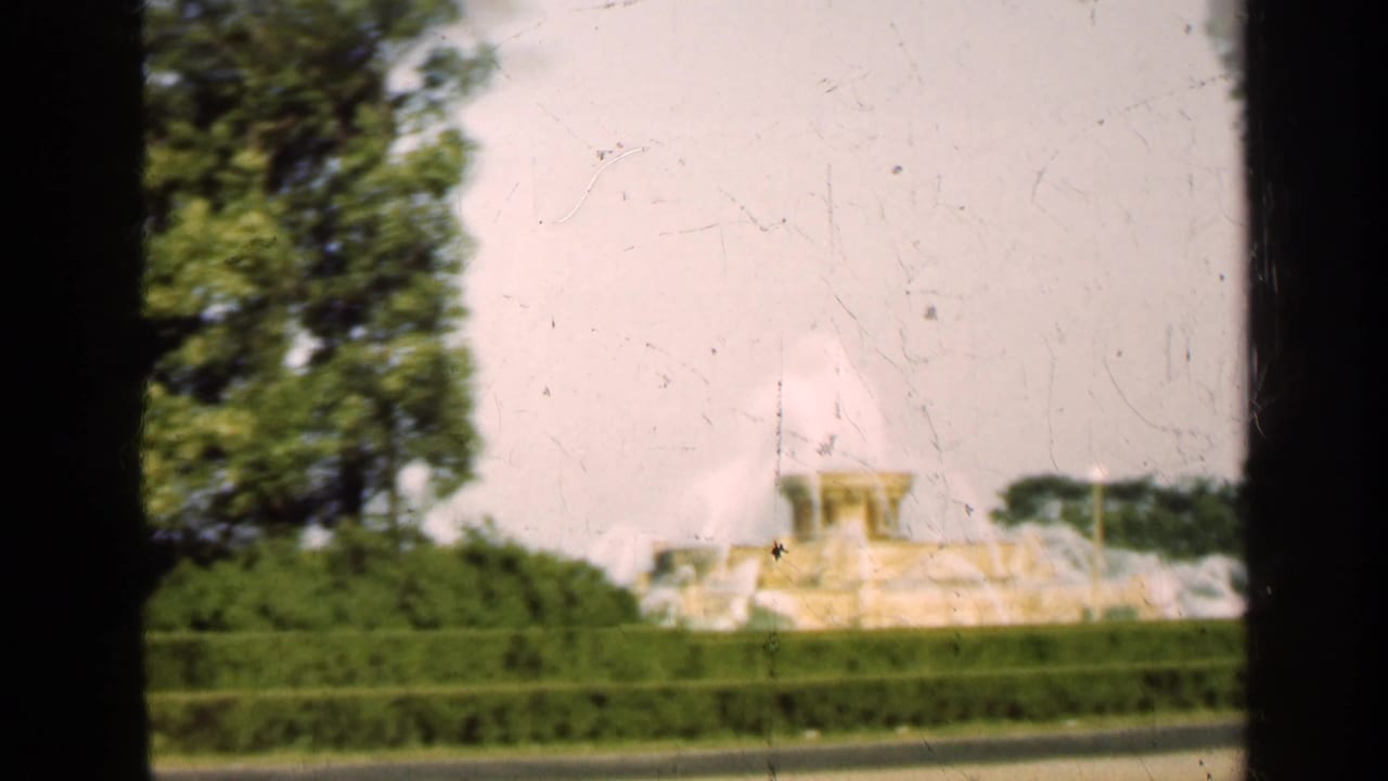 Distant fountain behind hedges and trees, seen through dark vertical frame edges.