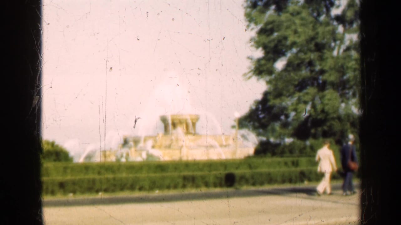 Two pedestrians walk along a paved path with a fountain and hedges behind them.