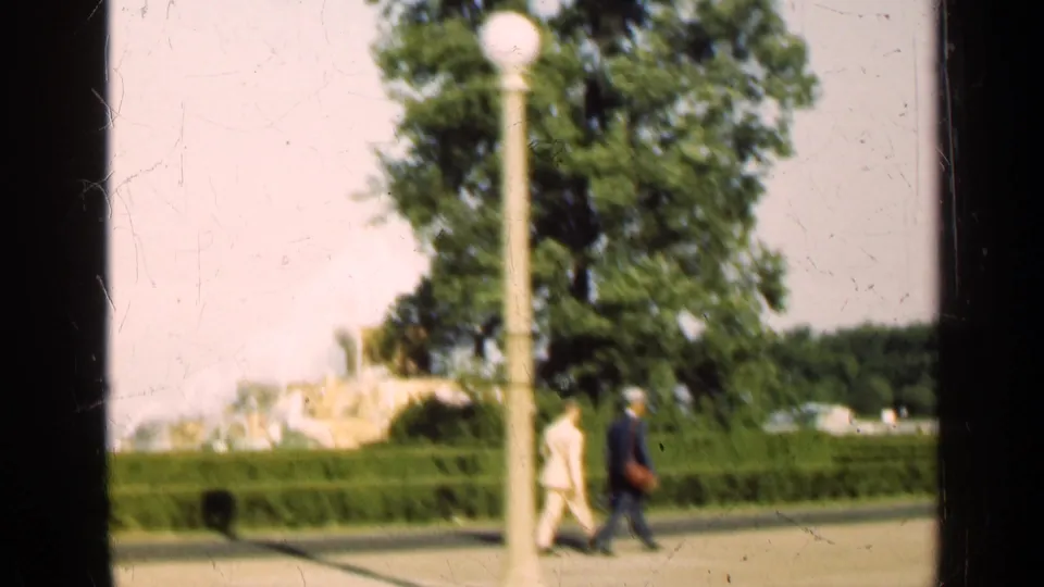 A globe lamp and two pedestrians cross the foreground of a 1947 Illinois park view with hedges, trees, and fountain spray behind.
