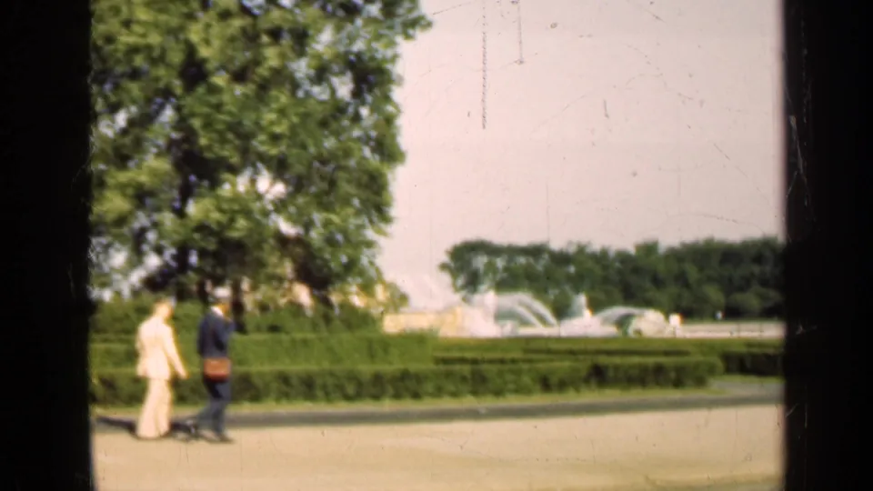 Two visitors continue along the path in the 1947 Illinois park footage, framed by hedges, trees, fountain spray, and dark film edges.