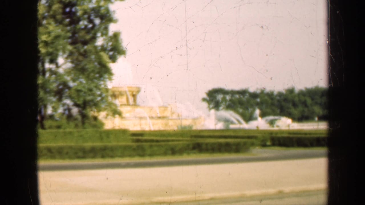 1947 Illinois 8mm frame showing a distant ornate fountain with water jets, hedges, pavement, and trees at the edge.