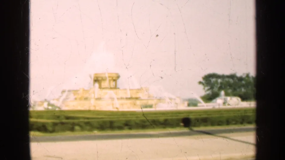1947 Illinois 8mm frame showing the fountain across a path, formal hedges, trees, and a small dark passerby in the foreground.