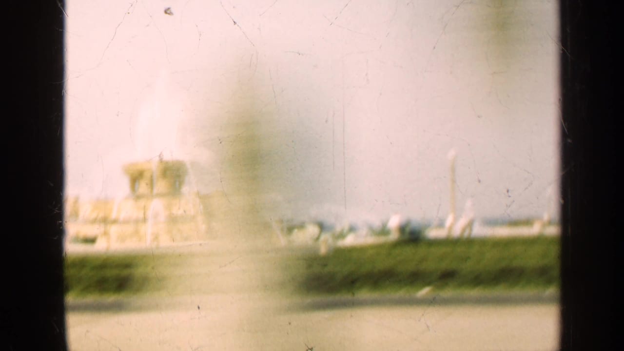 Blurred view of a fountain and lawn seen past a dark vertical divider.