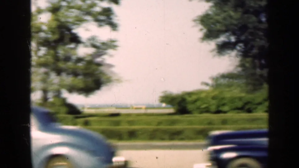 1947 Illinois 8mm frame looking across lawns and trees from a moving car, with two mid-century automobiles crossing the foreground.