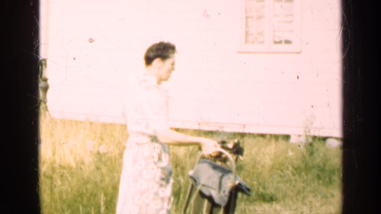Woman in a patterned dress stands near a black dog and bucket in a grassy yard.
