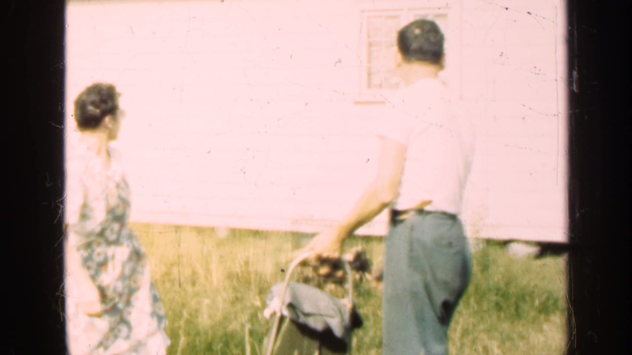 Man and woman stand near a black dog and bucket outside a light-colored house.