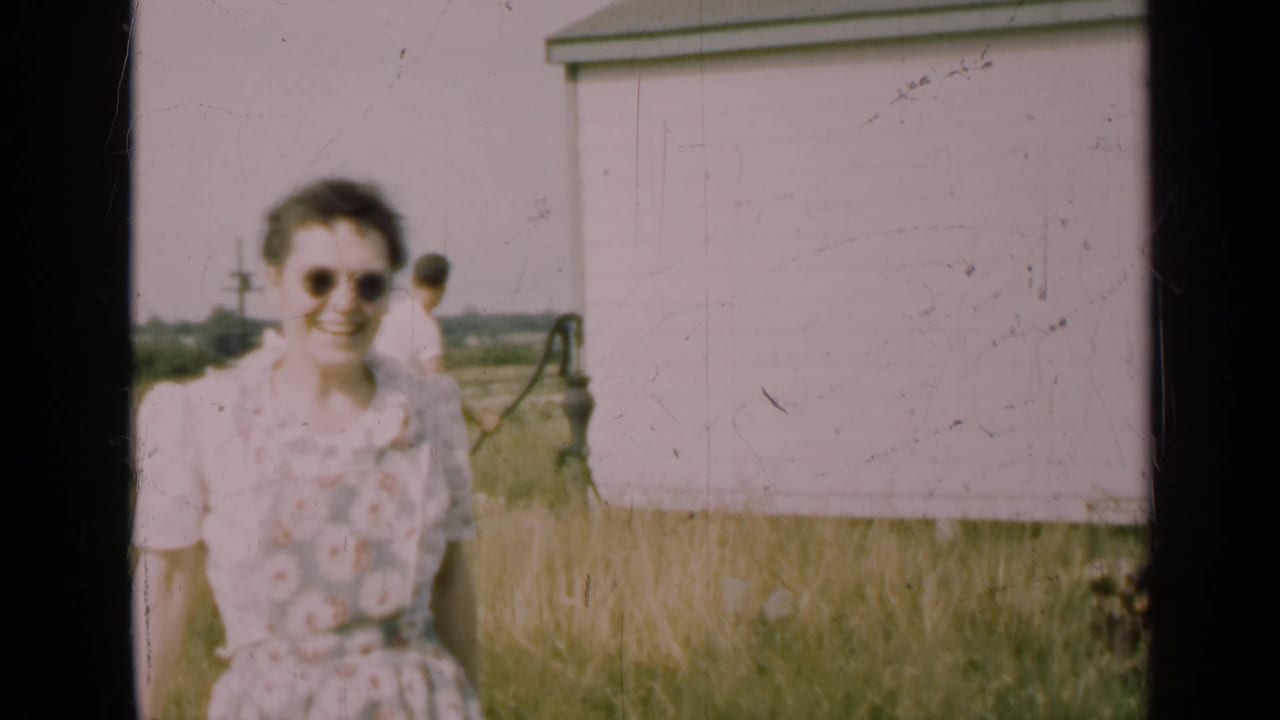 Woman in sunglasses smiles near a white farm building with a man and hand pump behind her.