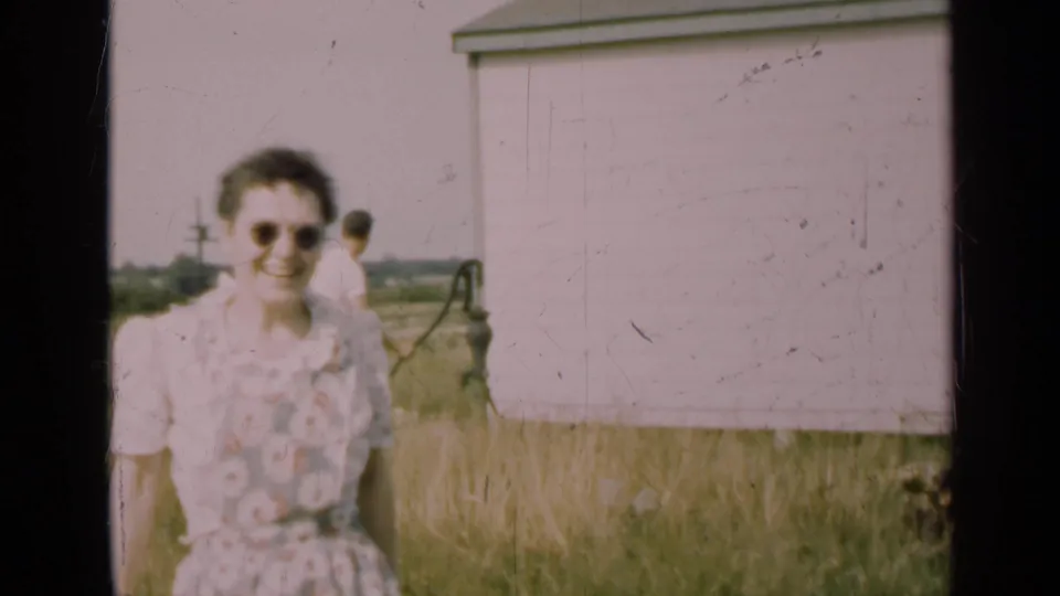 In 1947 Illinois farm footage, a woman in sunglasses and a floral dress smiles near a white shed while a man stands by a hand pump.