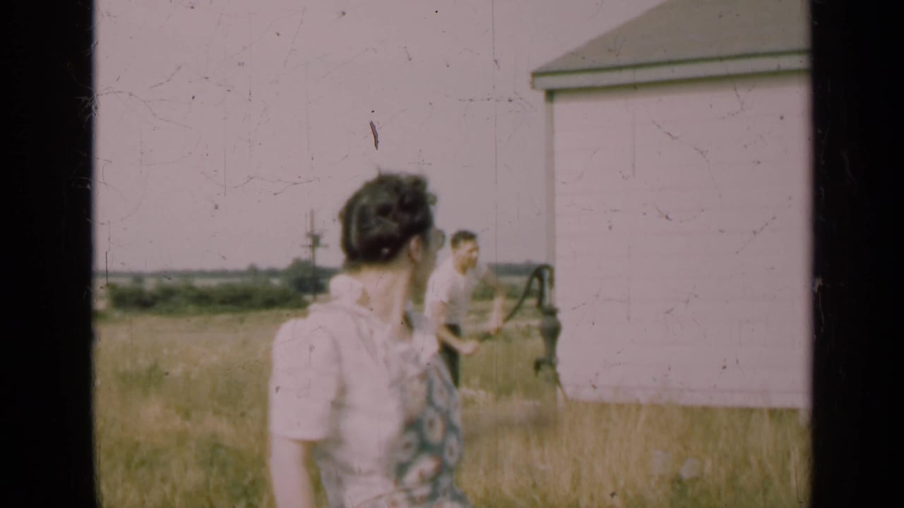 Woman turns toward a man working near a hand pump beside a white shed.