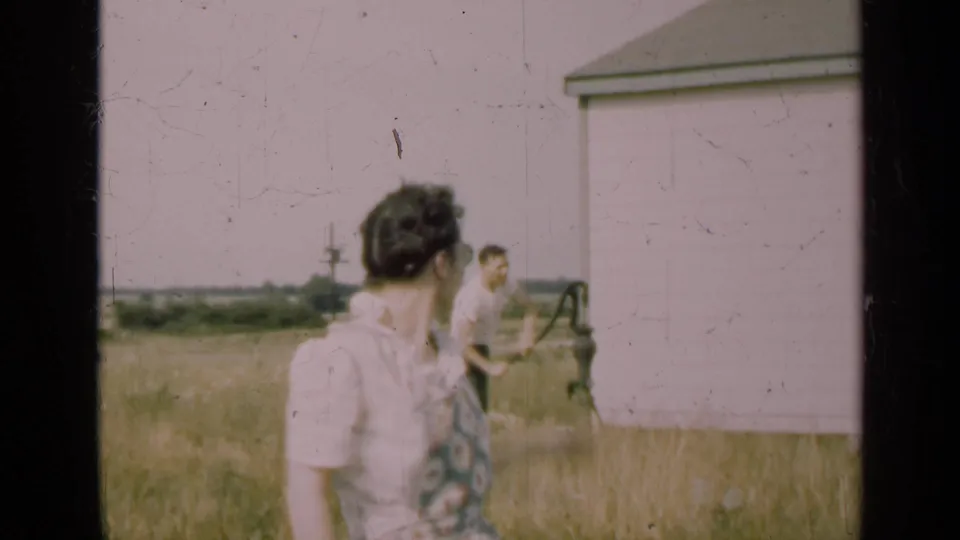 In the Illinois farm scene, the woman turns away from camera toward a man at a hand pump in tall grass beside a white shed.