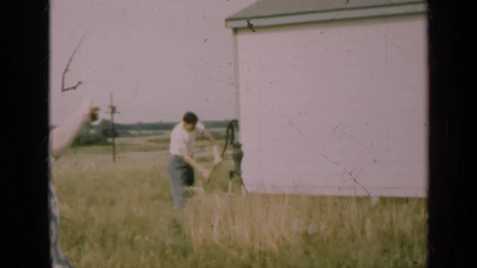 A man bends over the hand pump in tall grass as the woman enters from the left edge, gesturing across the farmyard.