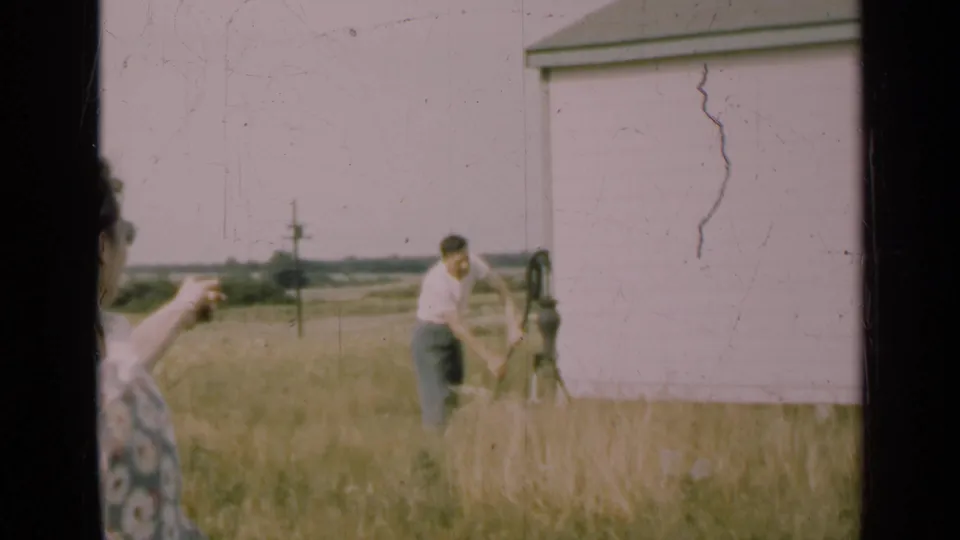 The woman points across the 1947 Illinois farm scene toward a man operating a hand pump near a white shed in tall grass.