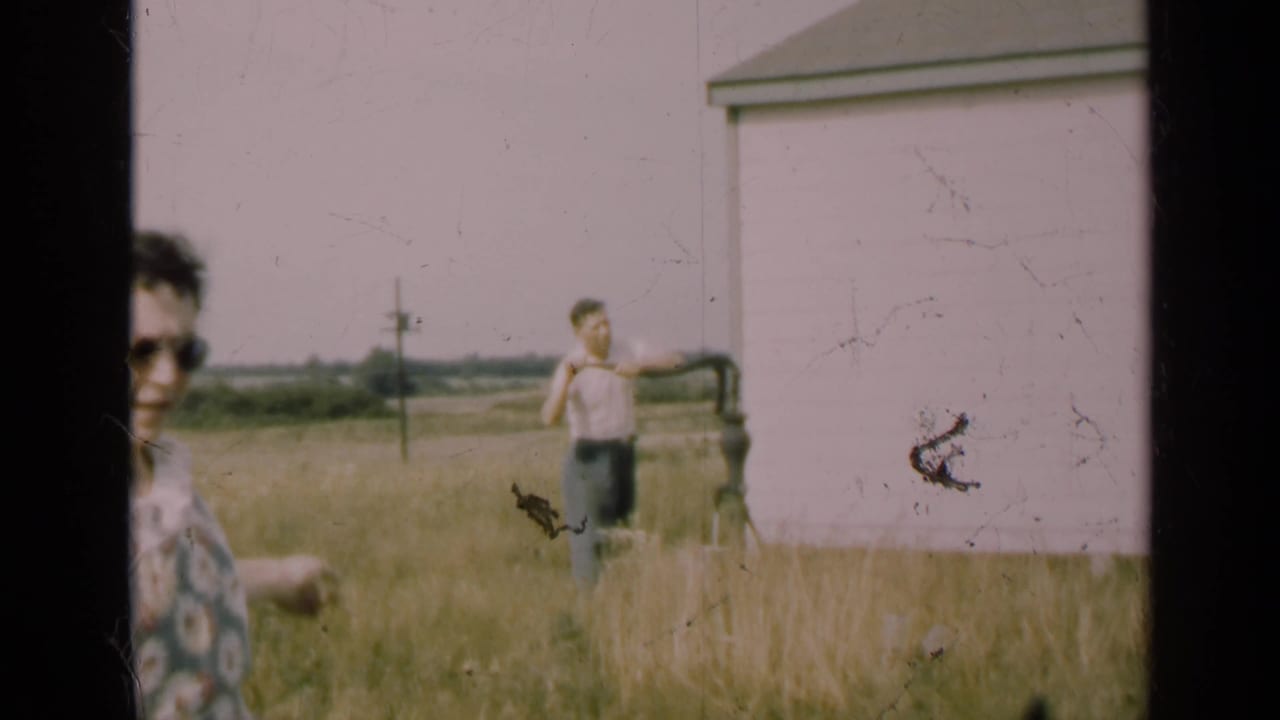 1947 Illinois home-movie frame showing a man at a hand-operated well pump near a rural house, with a partial foreground adult at left.