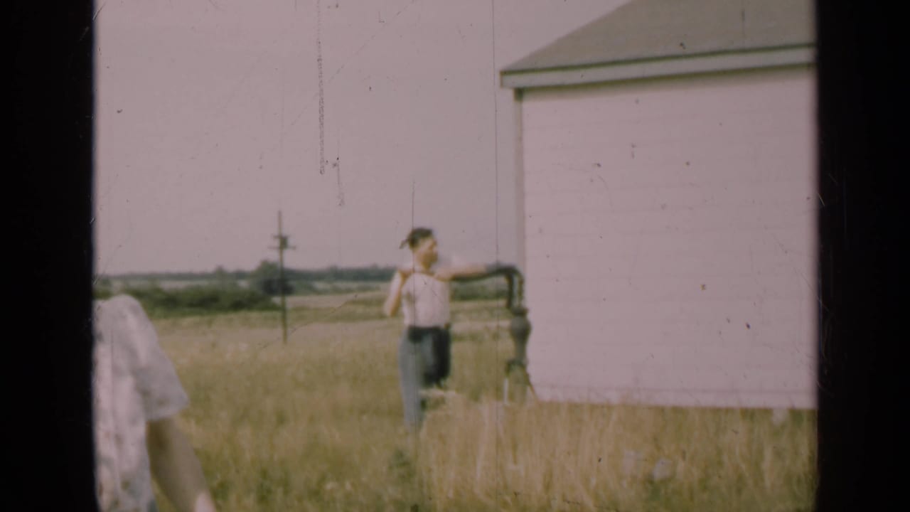 1947 Illinois rural footage shows a man operating a hand well pump next to a pale house wall, with open fields in the distance.