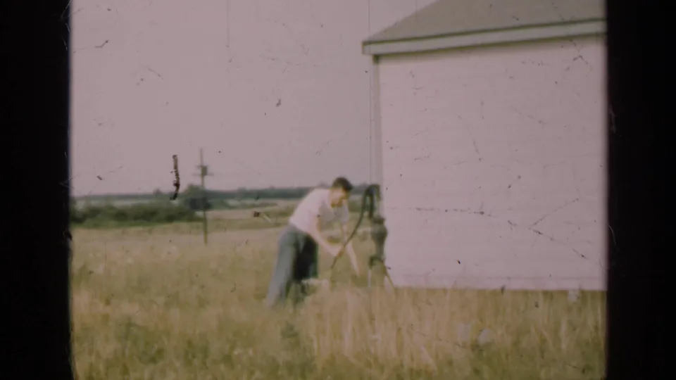 1947 Illinois home-movie frame showing a man bending at a hand pump by a rural house, with tall grass and fields beyond.