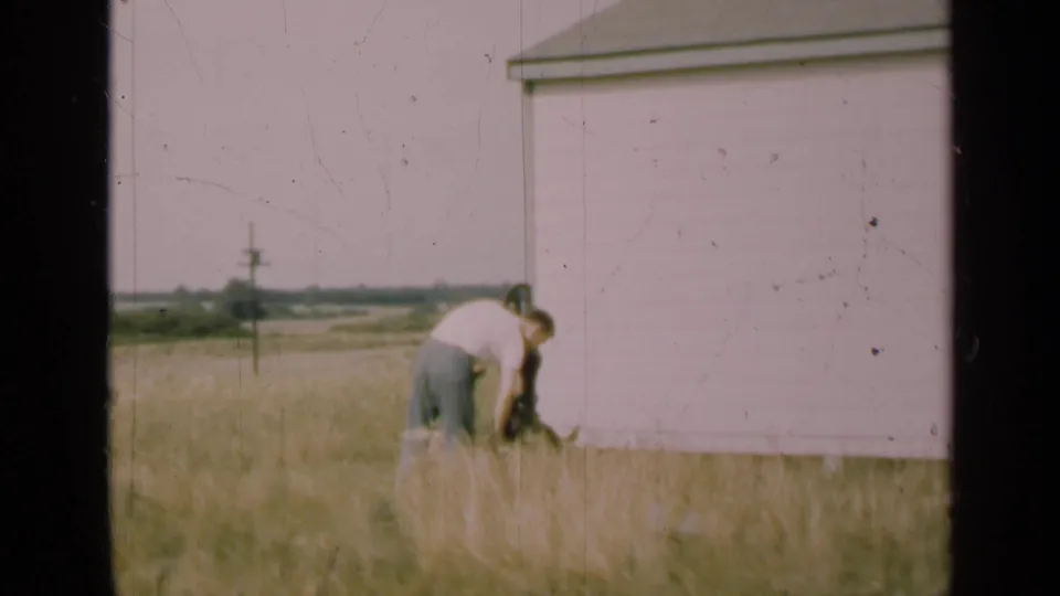 1947 Illinois rural scene with a man leaning over a hand-operated pump outside a pale house, filmed on scratched 8mm stock.