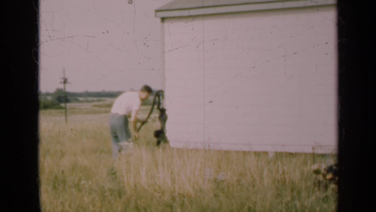 In 1947 Illinois home-movie footage, a man in a white shirt works near a hand pump by a white barn as a dog waits in tall grass.