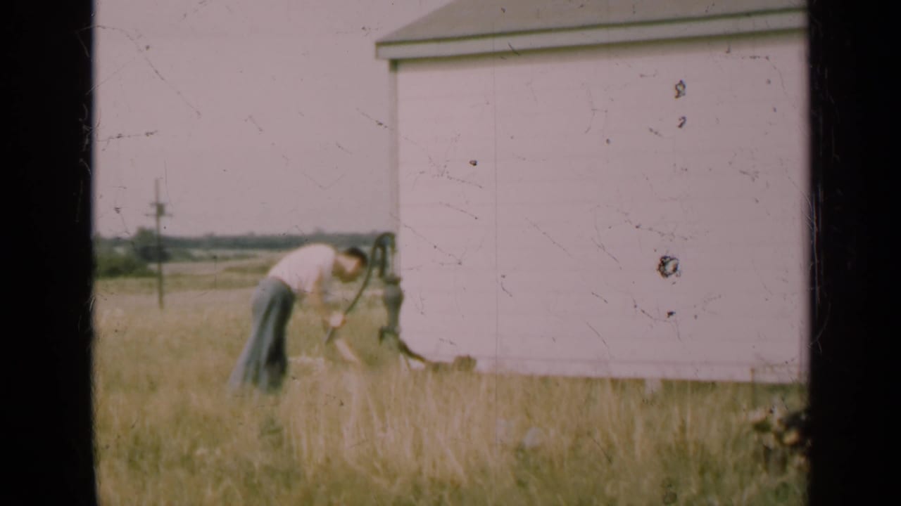 Man pumps water beside a white barn with a dog near the pump in tall grass.