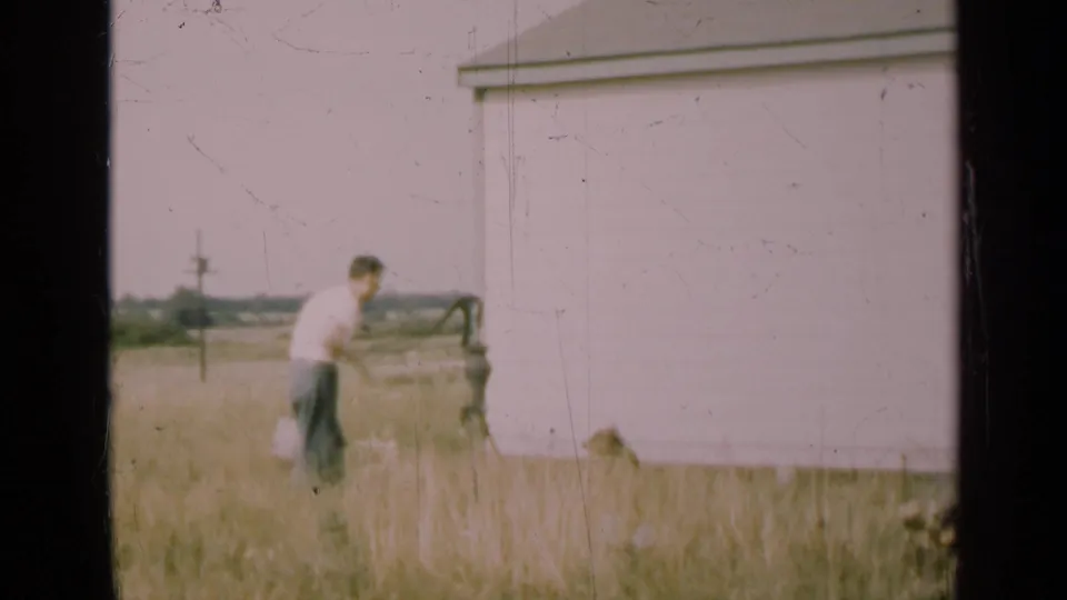 A man pauses beside the hand pump while the dog shifts away through the grass near the white barn in the 1947 Illinois scene.