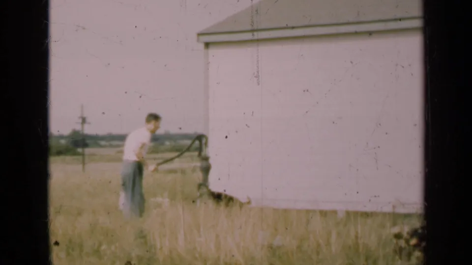 In the final frame, the man faces the hand pump beside the barn while the dog is partly low in the tall grass nearby.