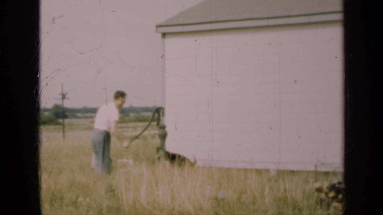 Man in a white shirt operates an outdoor hand pump beside a pale building in tall grass.