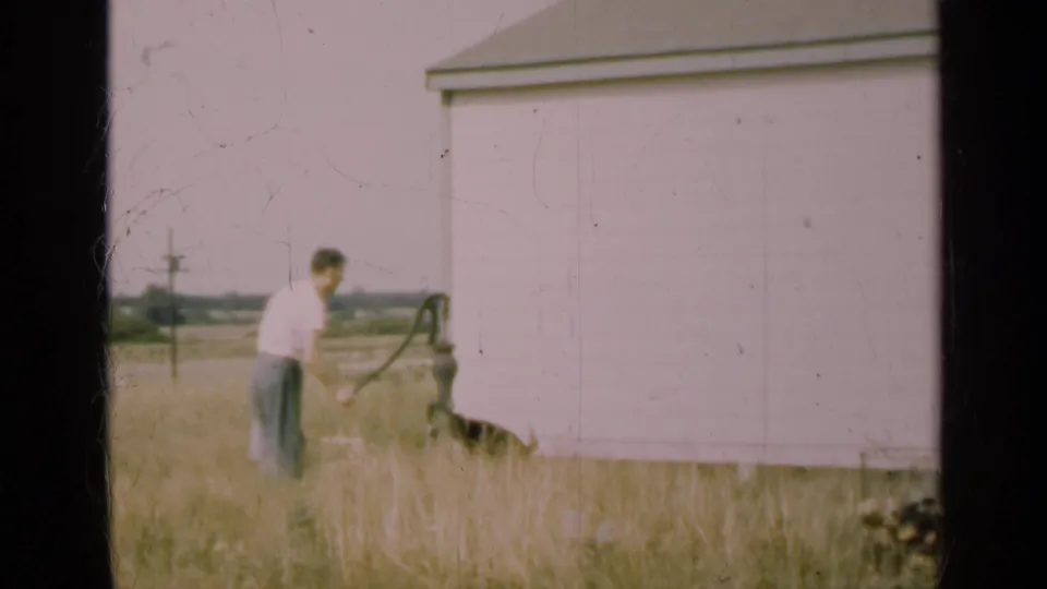 In 1947 Illinois home-movie footage, a man in a white shirt stands by a hand water pump near a pale rural outbuilding.