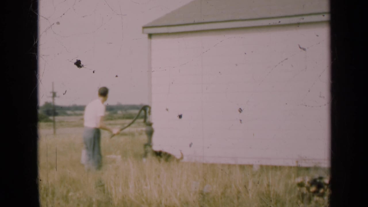 Man continues pumping water near a pale outbuilding with tall grass in the foreground.