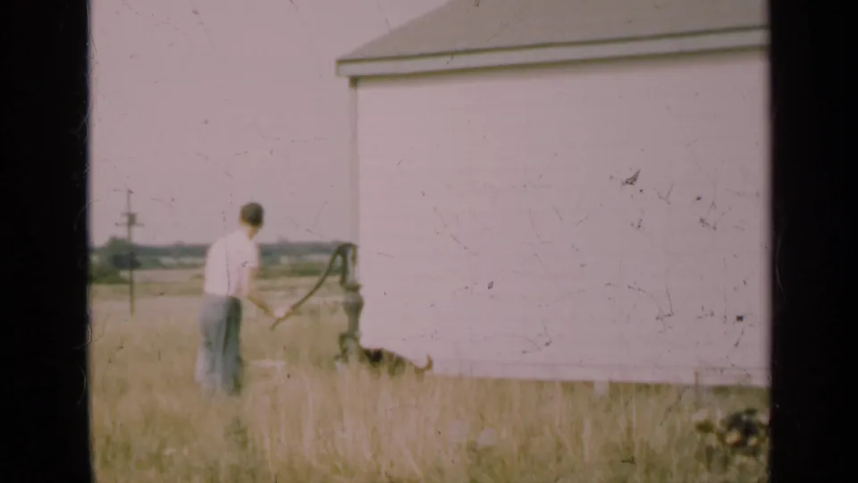 A soft 8mm frame shows the man beside the pump in tall grass, with the light-colored building occupying much of the background.