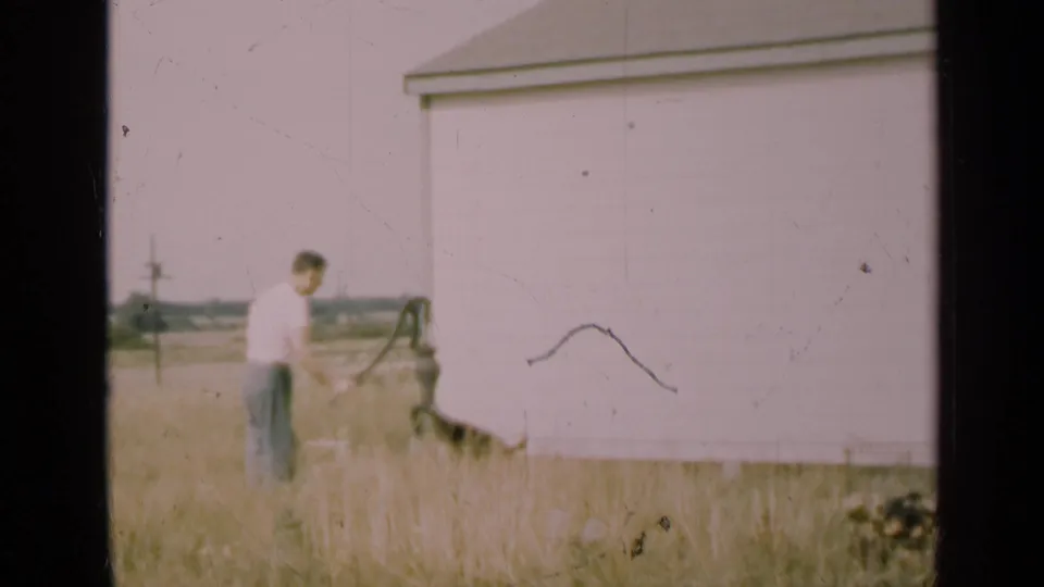 The final frame holds on the man and hand pump in 1947 Illinois, with faded color, scratches, and a pale rural outbuilding behind him.
