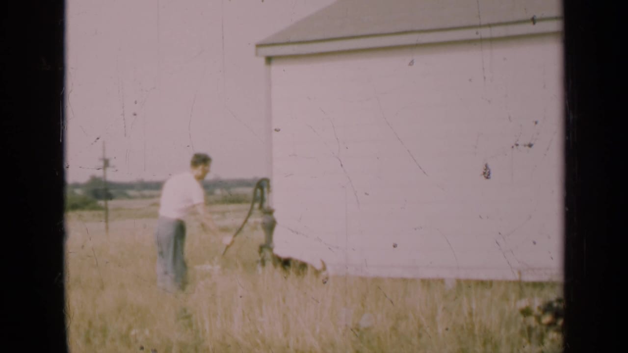 In 1947 Illinois home-movie footage, a man in a white shirt works a hand pump beside a white clapboard house and tall grass.