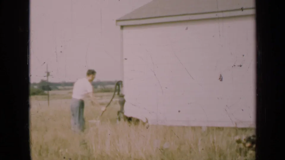 In 1947 Illinois home-movie footage, a man in a white shirt works a hand pump beside a white clapboard house and tall grass.