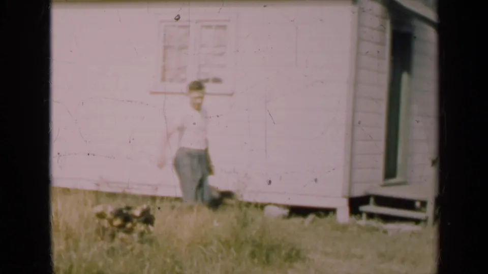 A man stands in tall grass near a small dog outside a white clapboard house, with a window and doorway visible in the frame.