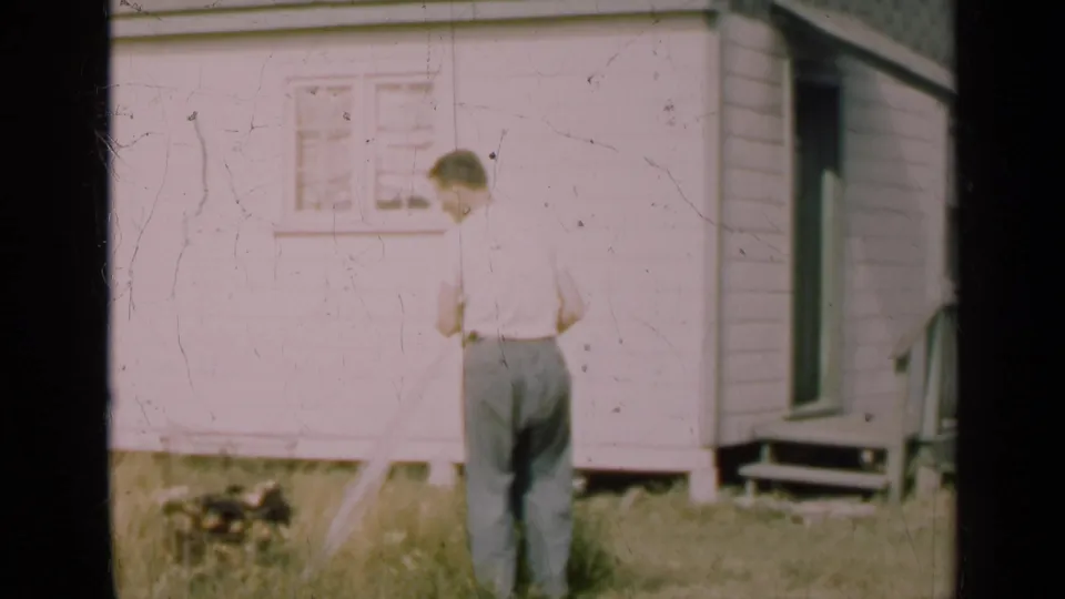 The man faces away in the grassy yard near the house, holding a narrow object as the doorway and window remain visible.