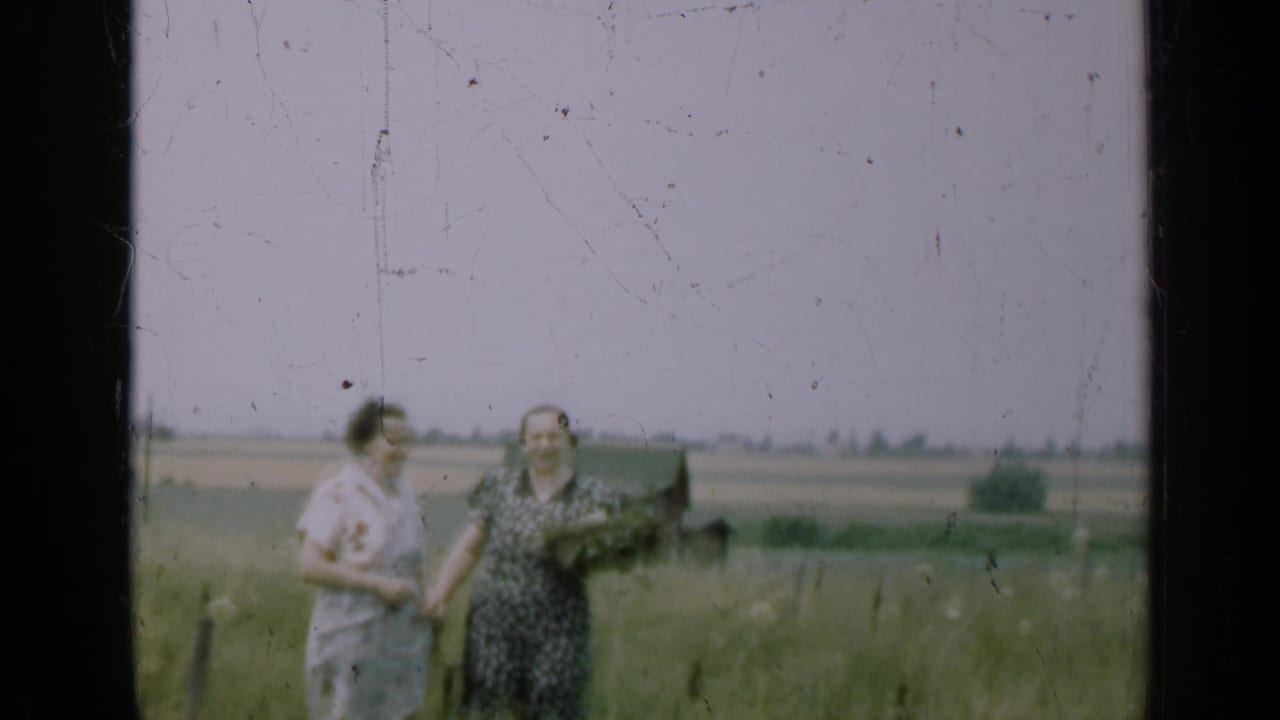 Two women in dresses stand in a grassy field with distant farmland behind them.