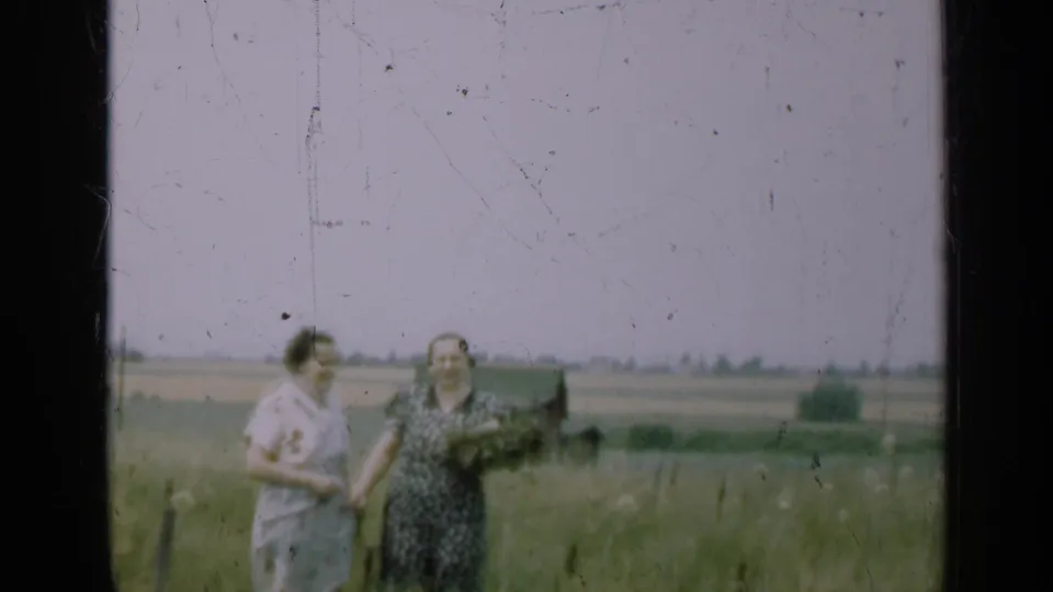 Two women in dresses stand together in a grassy Illinois field in 1947 home-movie footage, with soft focus and distant farmland visible.