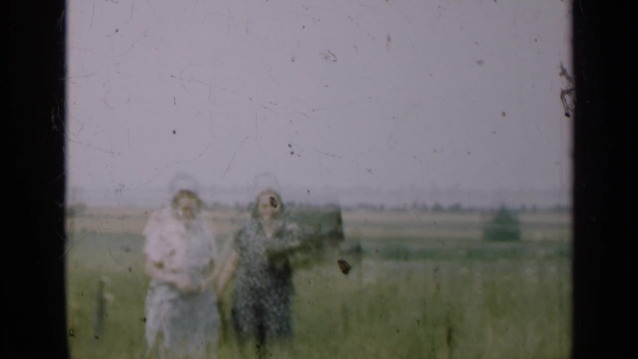 In the 1947 Illinois home-movie scene, two women stand in tall grass as one raises a hand near her face, with farmland in the distance.