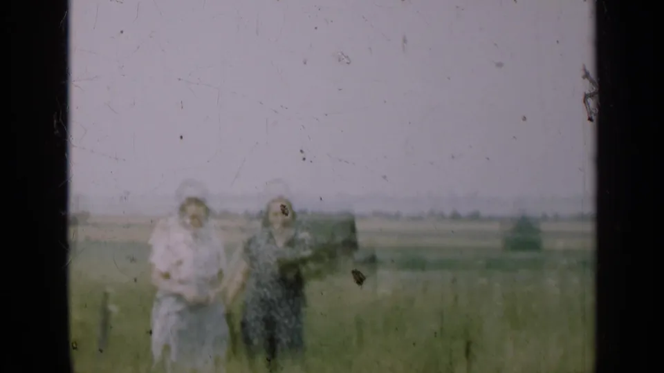 In the 1947 Illinois home-movie scene, two women stand in tall grass as one raises a hand near her face, with farmland in the distance.