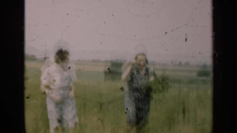 Two women stand apart in a rural Illinois field, one adjusting or touching her hair, in soft 1947 8mm home-movie footage.